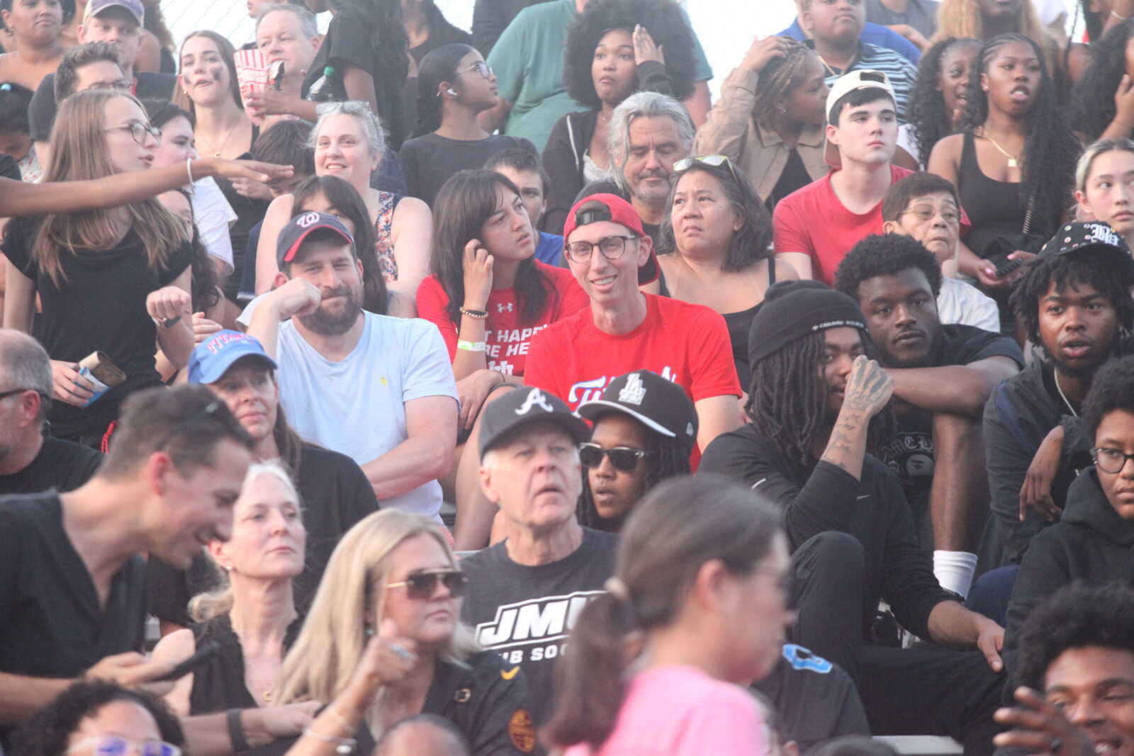 Large crowd of diverse people seated and standing at an outdoor event, some wearing caps and sunglasses, looking in various directions.