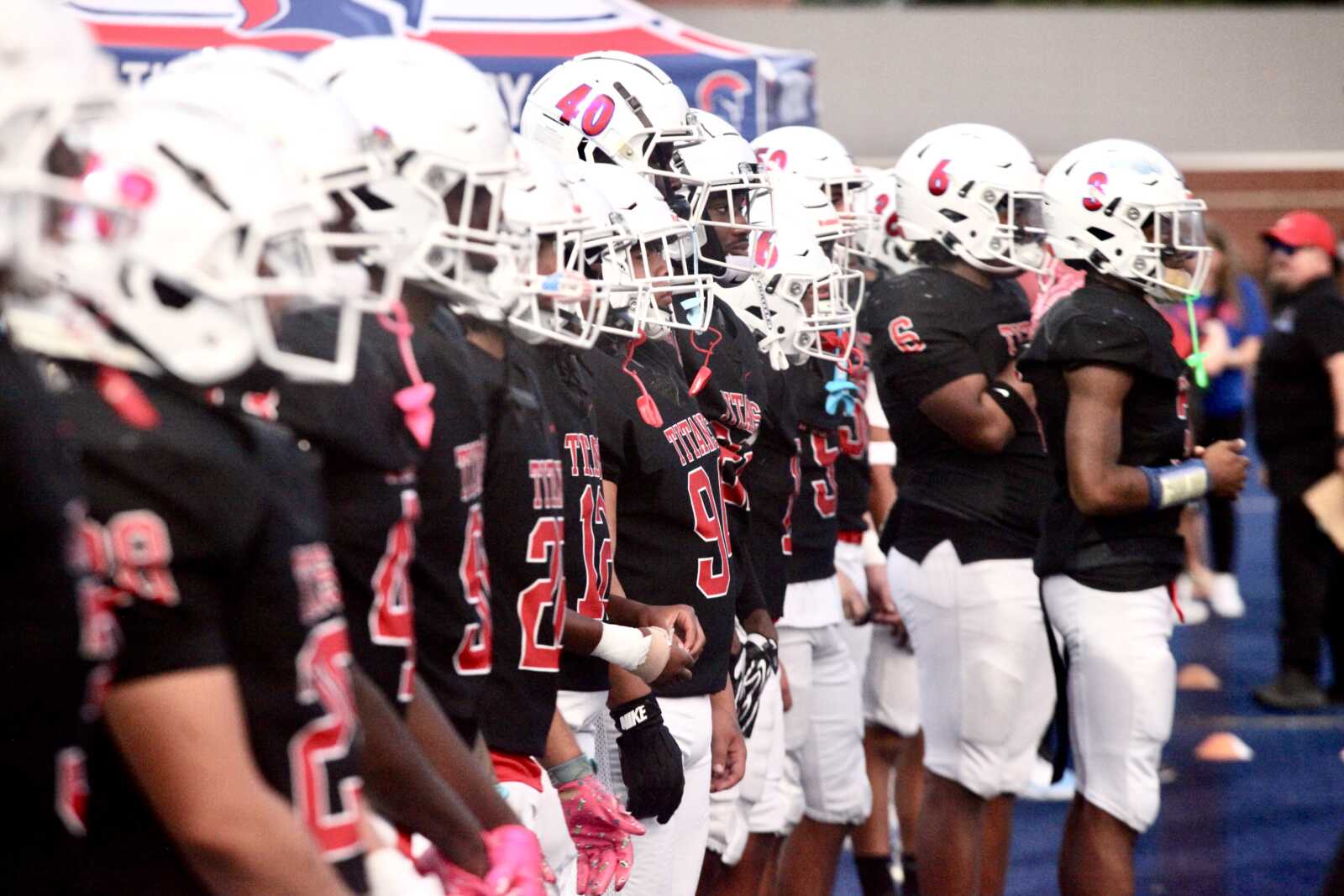 Line of football players in black jerseys and white helmets stand in a row before a game, hands at their sides.