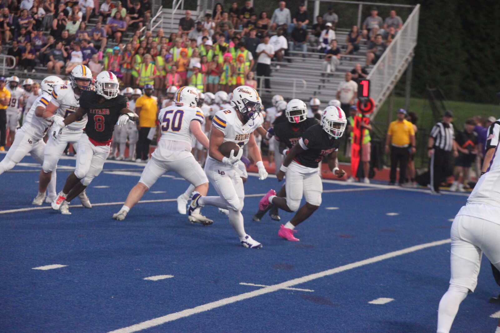 A football running back in a white uniform carries the ball while defenders in black chase him on a blue turf field with a crowded stands in the background.