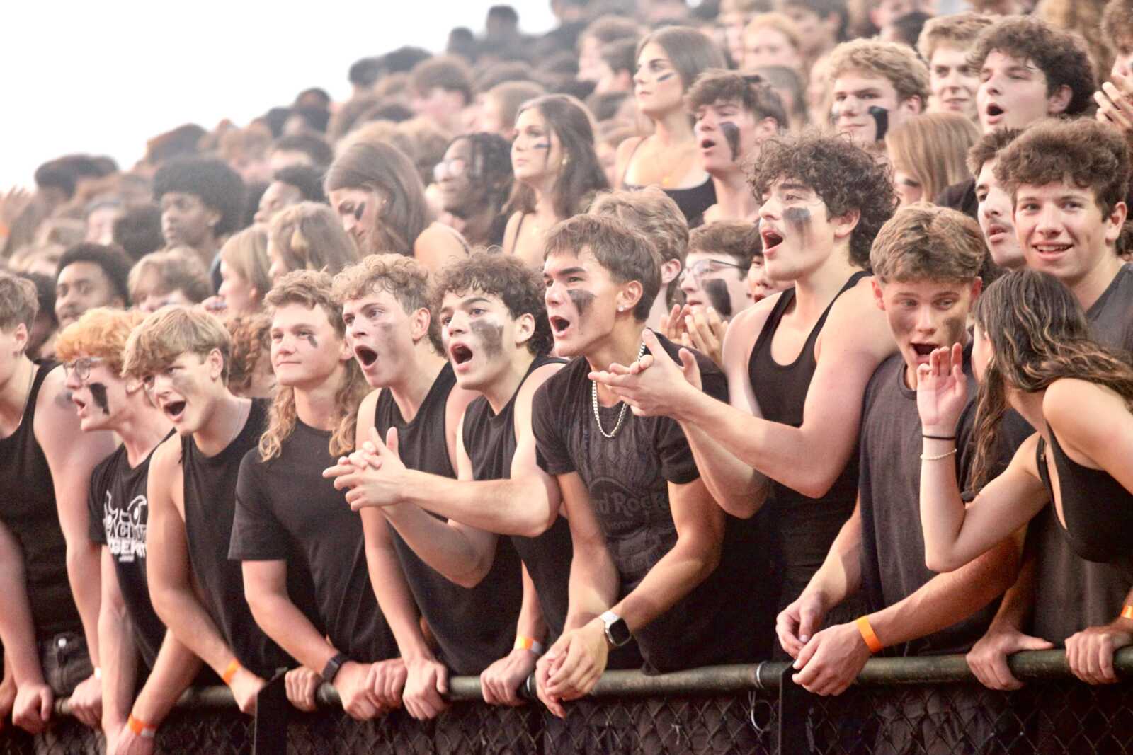 Large crowd of young people cheering behind a barrier at a stadium or event, many with black face paint and tank tops.
