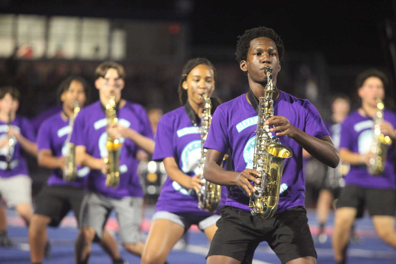 Young man in a purple shirt playing a saxophone on a football field during a band performance, with other players in purple shirts in the background.