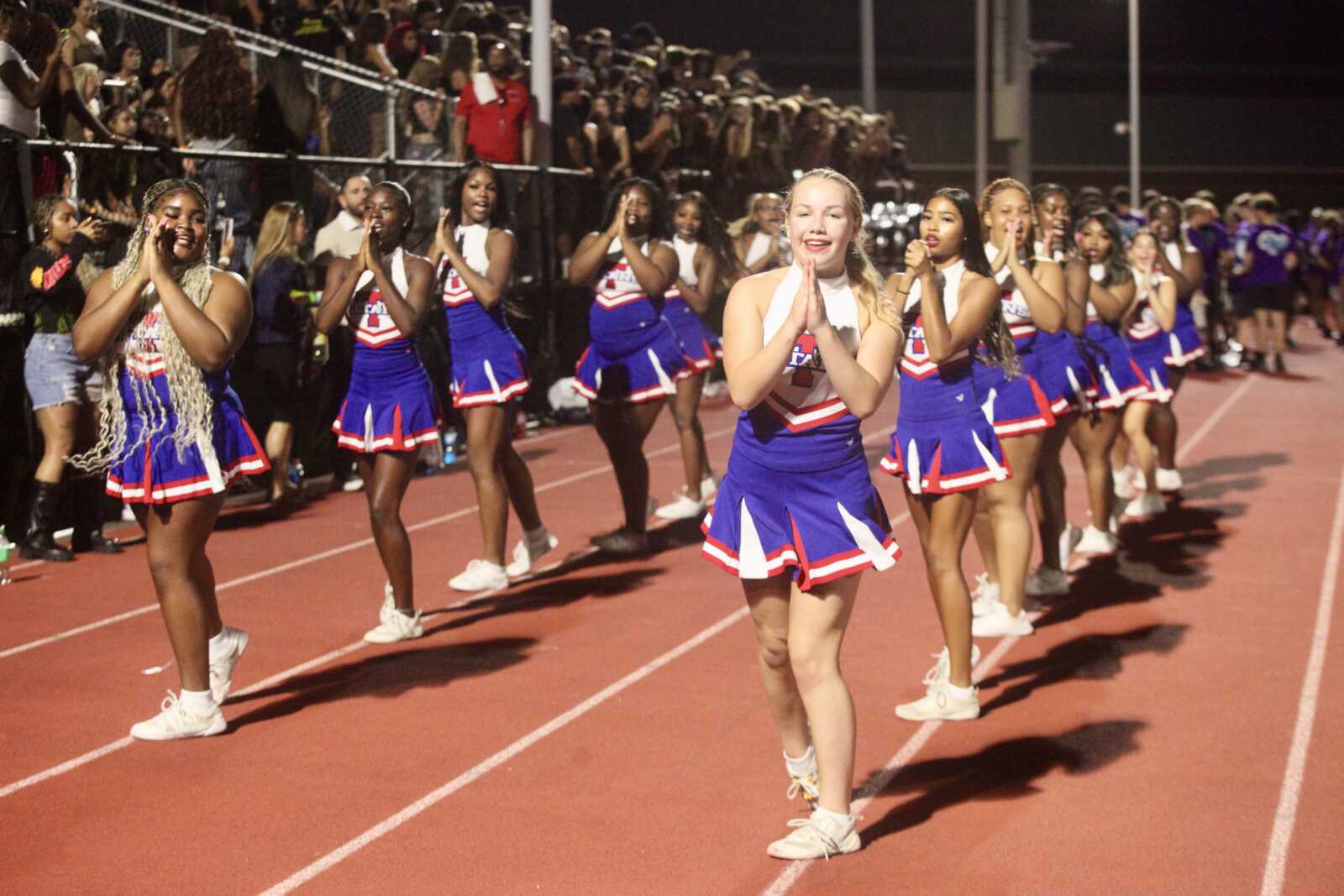 Line of cheerleaders in blue, white, and red uniforms performing on a track as a nighttime crowd watches from the stands.