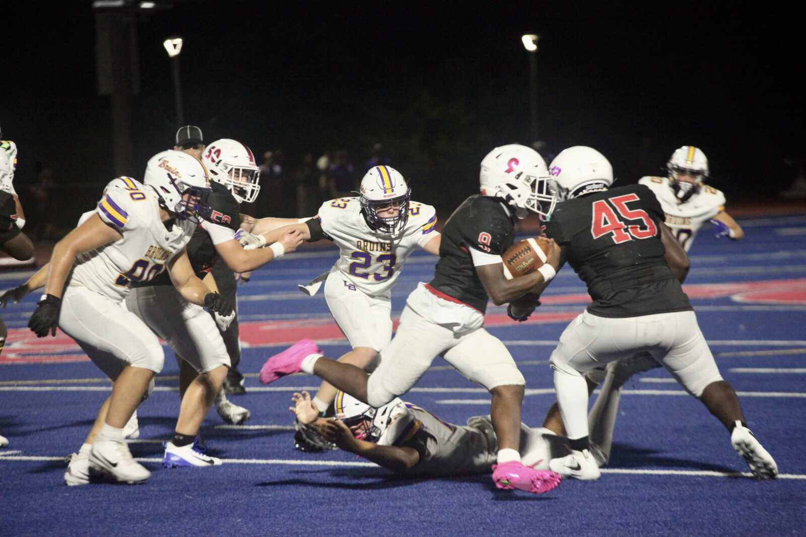 Football players collide on a blue turf field at night; a Black-jersey runner holds the ball as defenders close in.