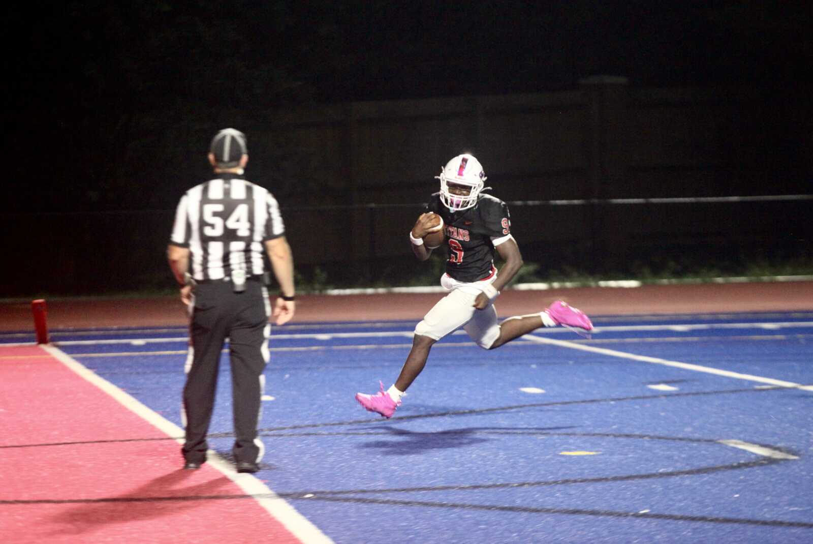Night football scene: a player in a black jersey, white pants, and pink cleats runs with the ball toward the end zone while a striped-referee watches from the sideline on a blue field reconnecting the end zone.