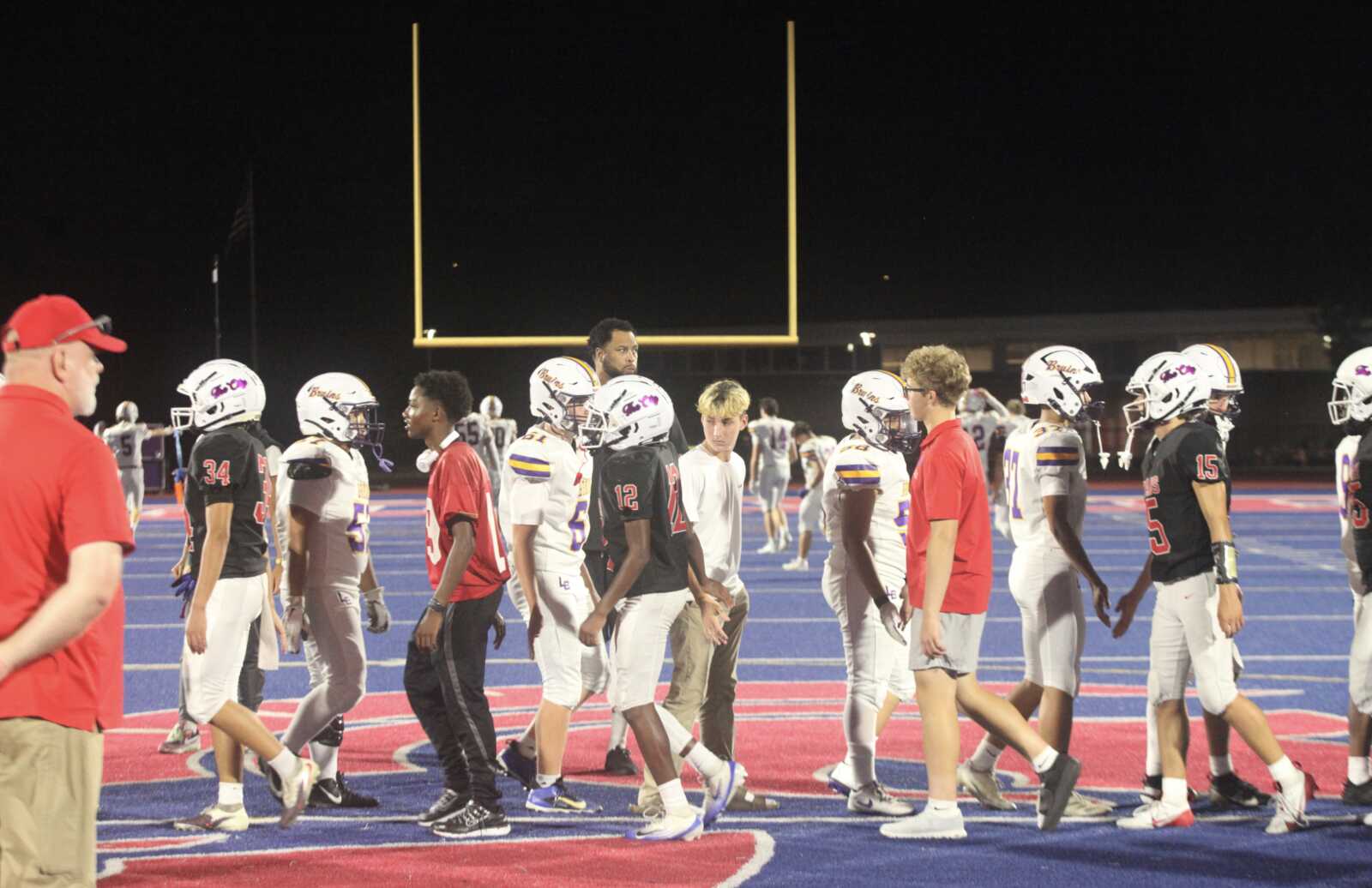 Two football teams shake hands along a blue end zone after a night game, coaches in red shirts nearby.