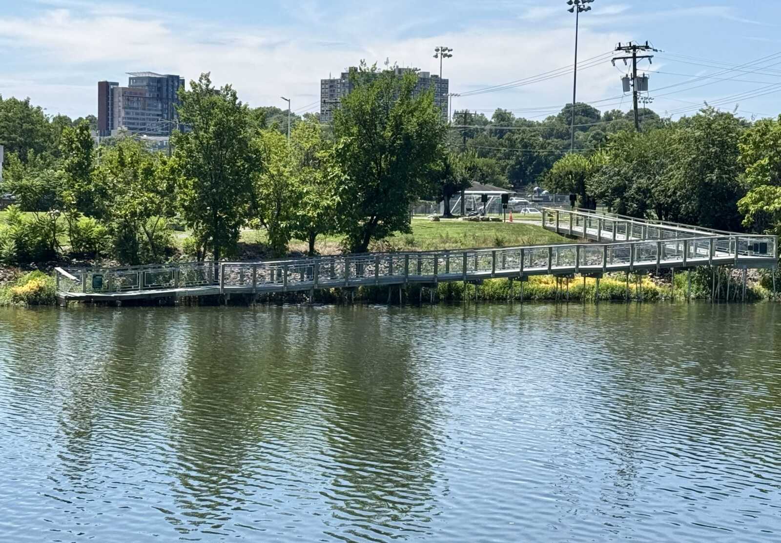 Long metal pedestrian bridge over a calm lake, bordered by trees and a grassy park with buildings in the background.