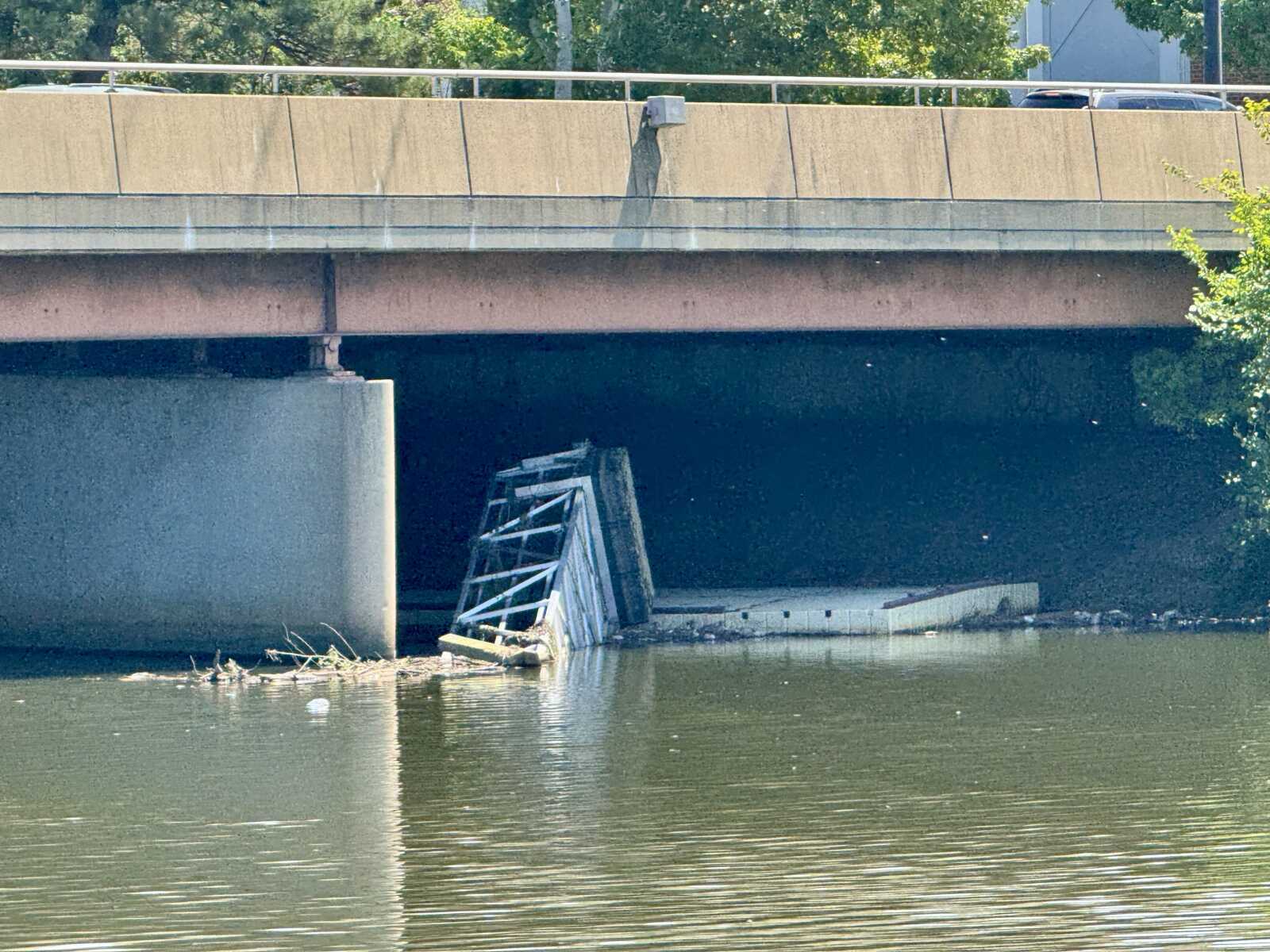 Under a concrete highway overpass above a river, a pile of rusted metal debris and broken fencing rests against the pier with water in the foreground.