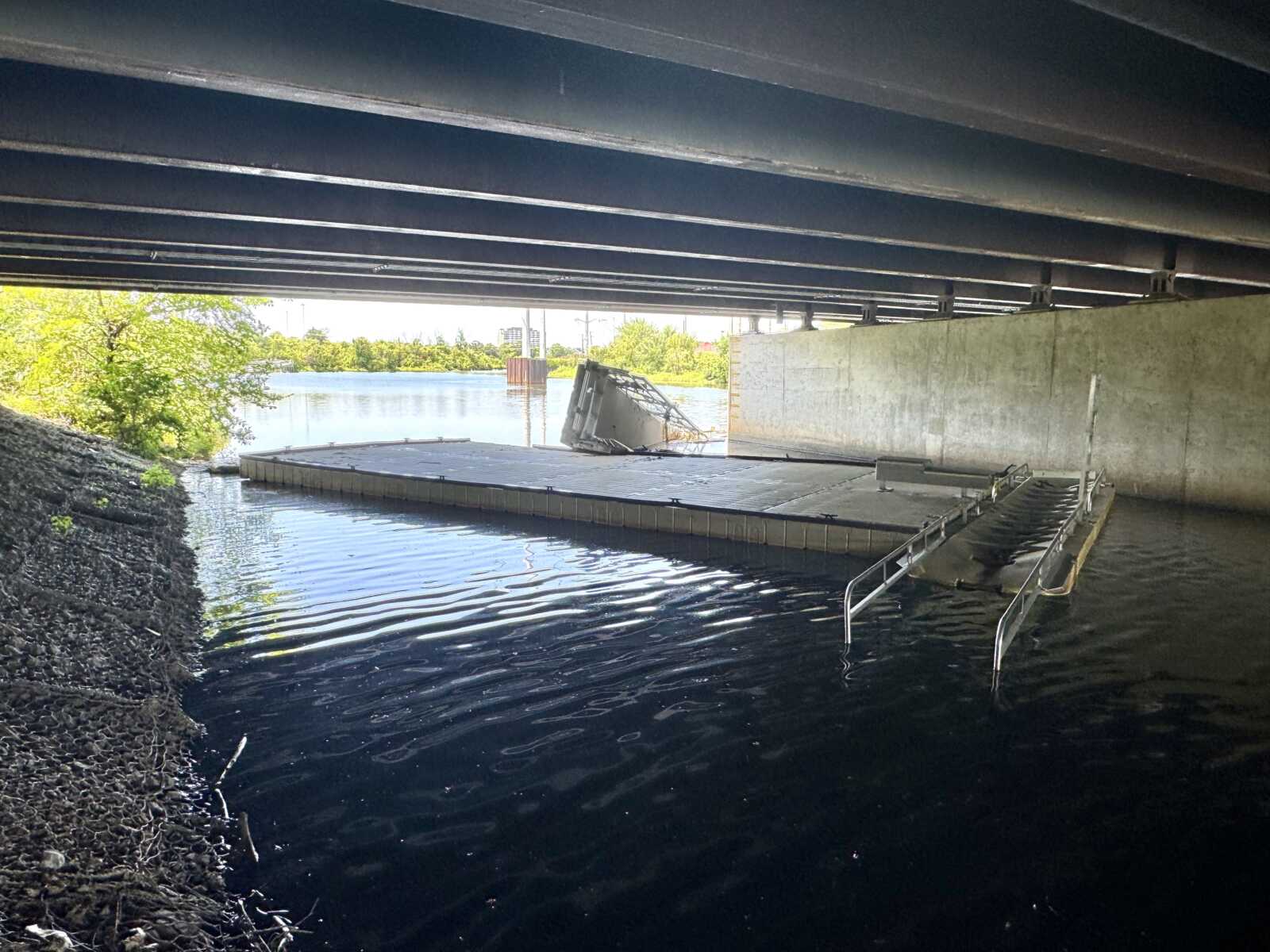 Under a bridge over a calm river, a floating platform with a toppled container and a metal ramp nearby.
