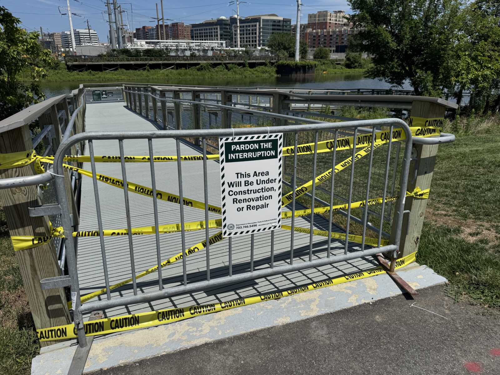 Construction site along a river with a fenced gate and bright yellow caution tape surrounding a boarded walkway