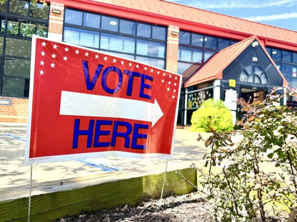 Red political sign reading 'VOTE HERE' with a white arrow, placed outdoors near a brick building entrance.