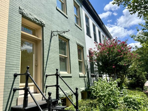 Green brick townhouse with a beige door, black railing, and a pink flowering bush by the path on a sunny day.