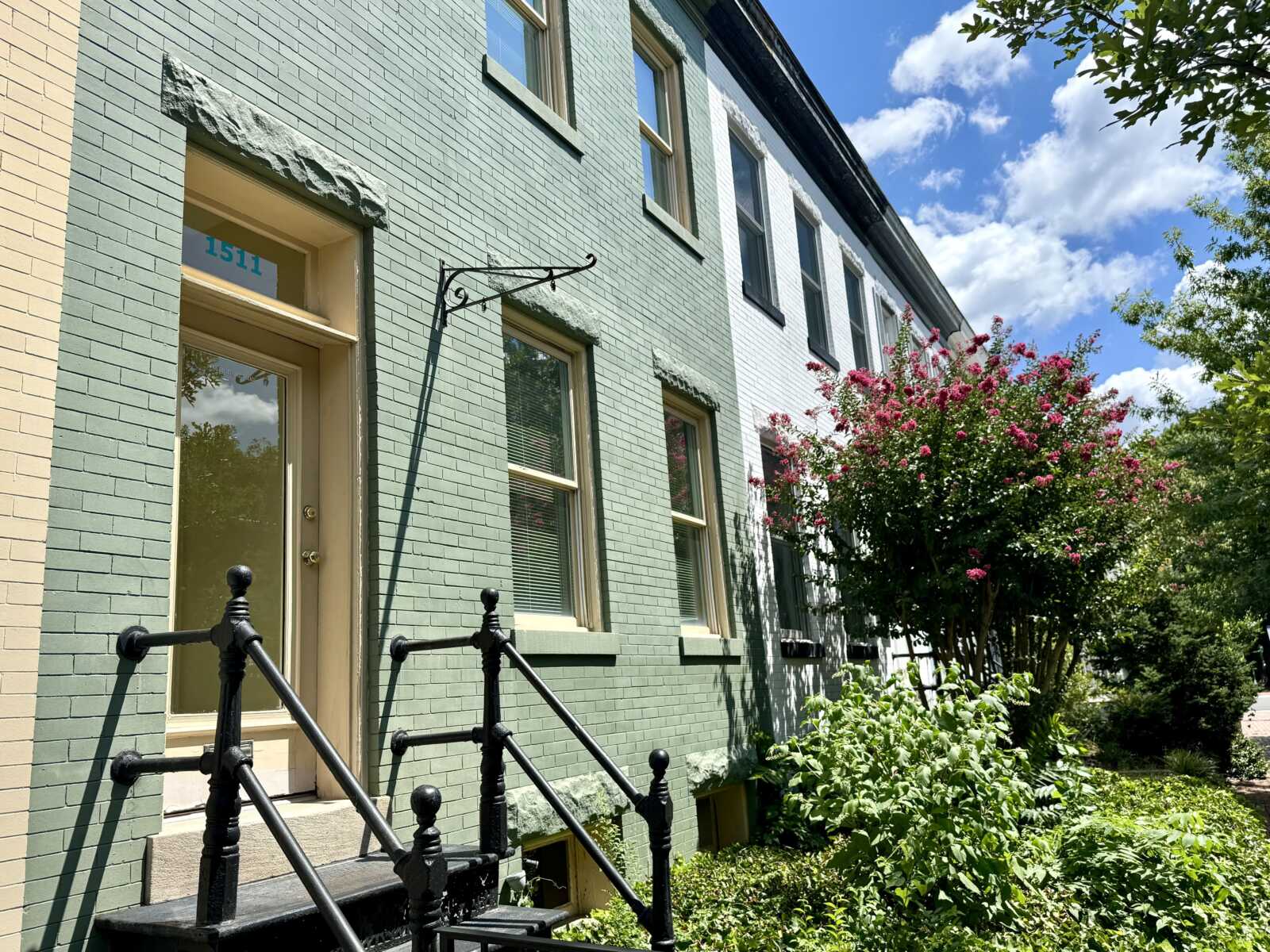 Green brick townhouse with a beige door, black railing, and a pink flowering bush by the path on a sunny day.