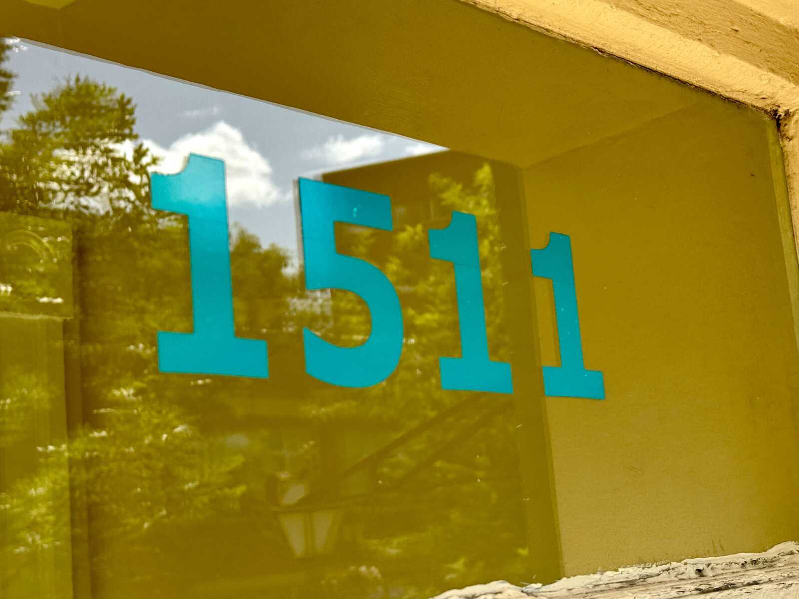 Turquoise address numbers '1511' on a reflective glass surface showing trees outside.