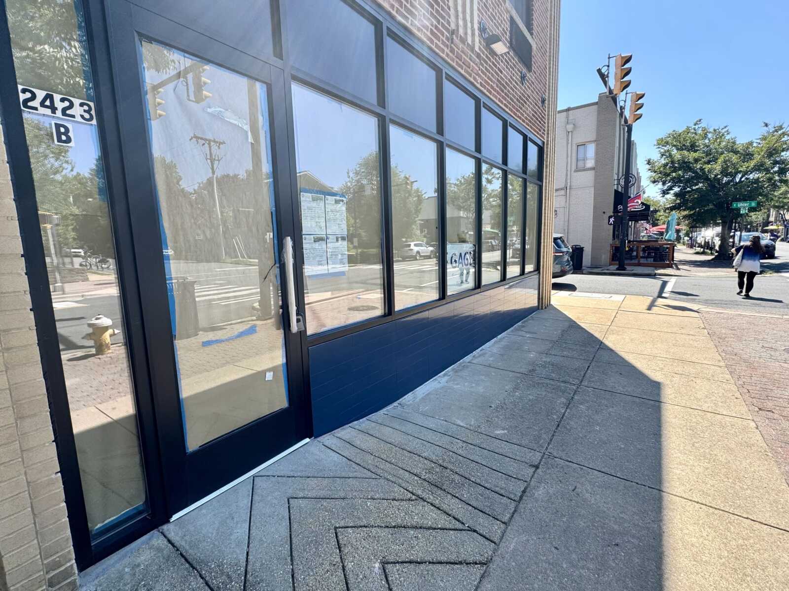 storefront with large glass windows along a brick building on a sunny street; a person walks away on the sidewalk.