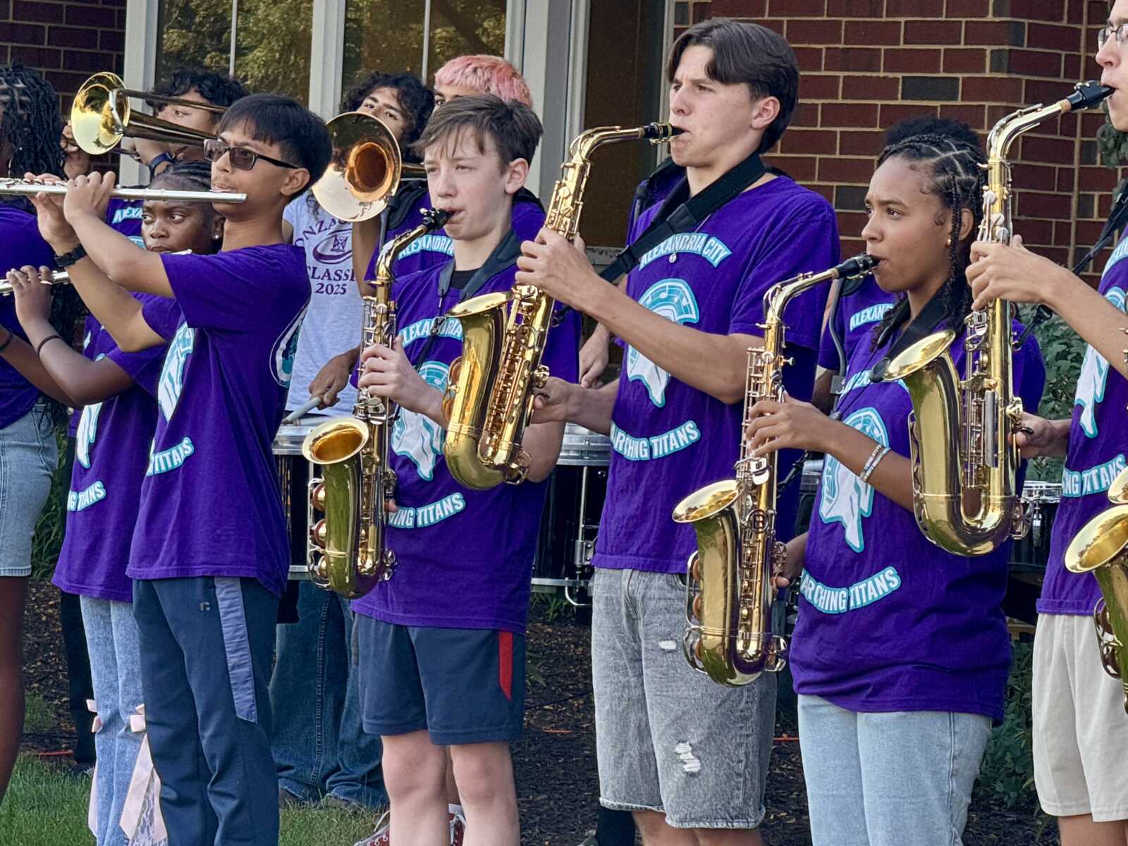 Group of young musicians in purple shirts playing trumpets and saxophones outdoors by a brick building.