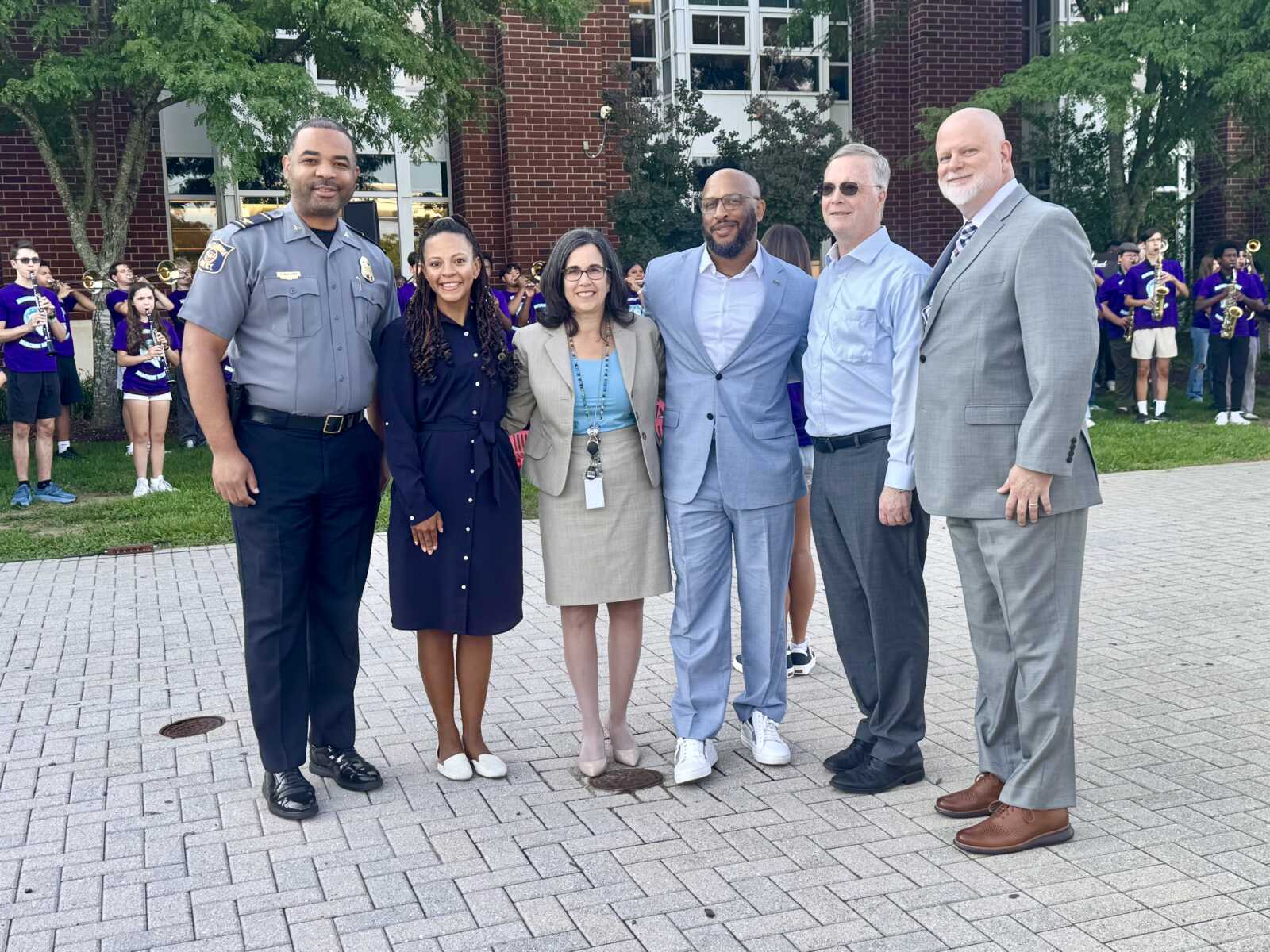 Six adults posing outdoors in front of a brick building, including a police officer, two women, and three men in suits; a marching band is seen in the background with purple shirts.