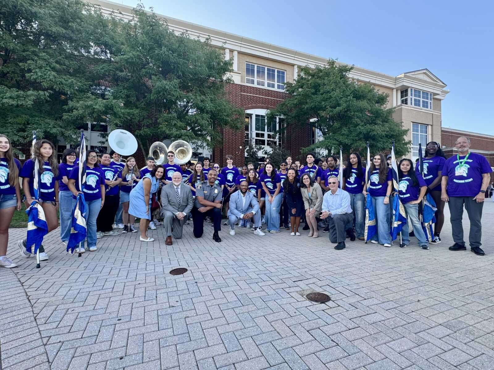 Large group of students in purple shirts posing outdoors with flags and brass instruments in front of a brick school building.
