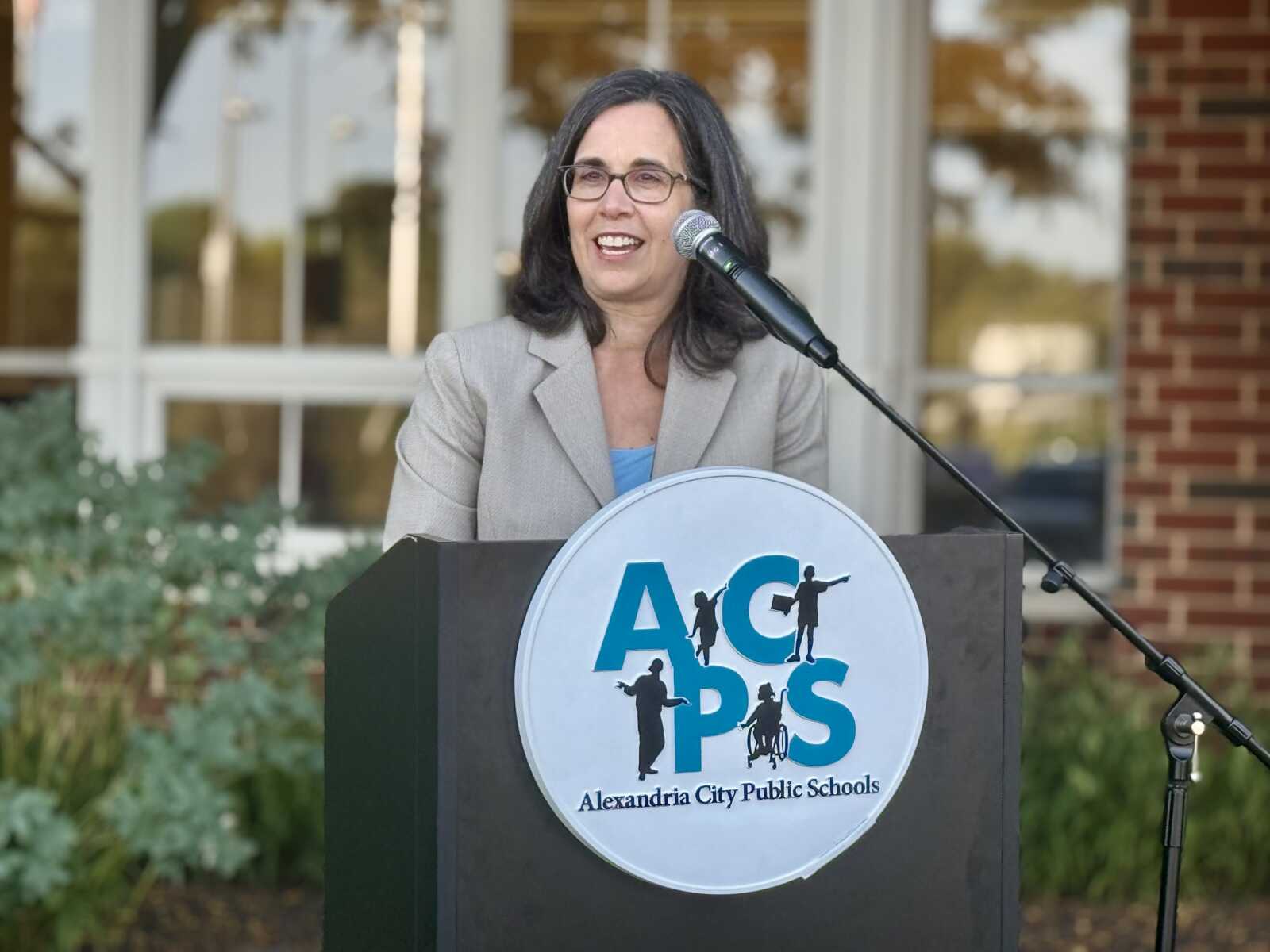 Woman in a gray blazer speaks at a podium with the AACPS logo outdoors.