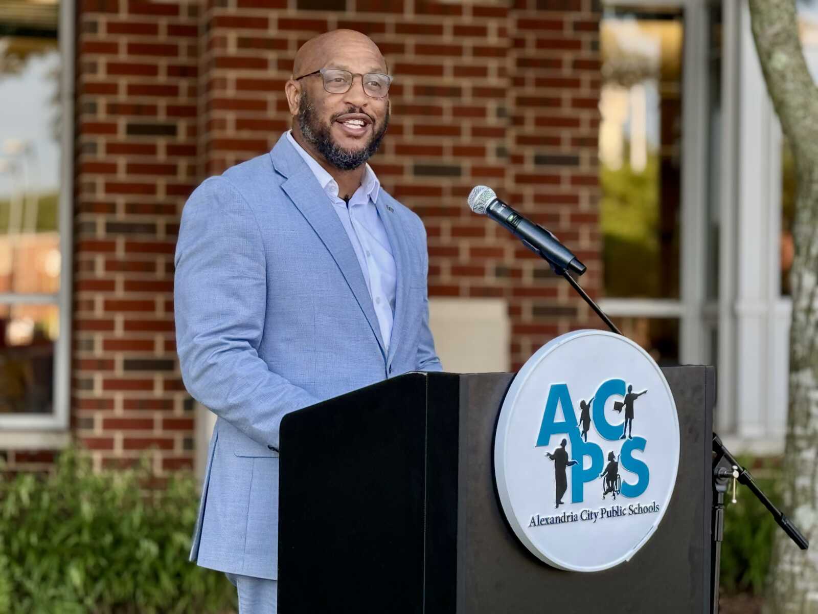 Man in a light blue suit giving a speech at a podium with a school district logo outdoors.