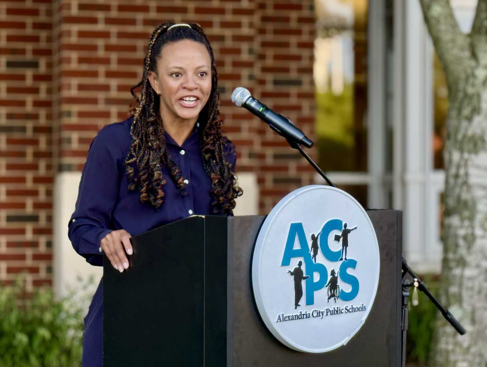 Woman with braided hair speaks at a podium with a microphone outdoors, brick building behind; AACPS logo on the podium.