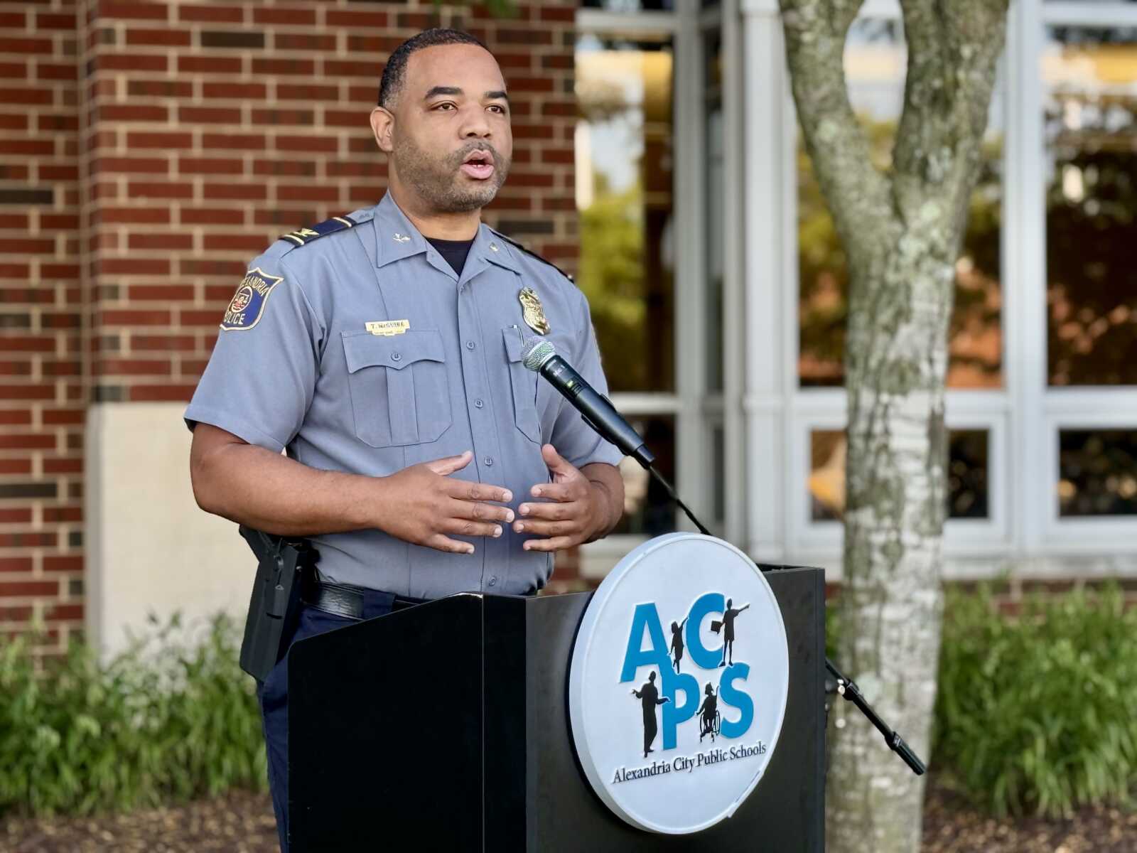 Police officer in uniform speaking at a podium with a microphone outside a brick building, likely a school, during a public address.