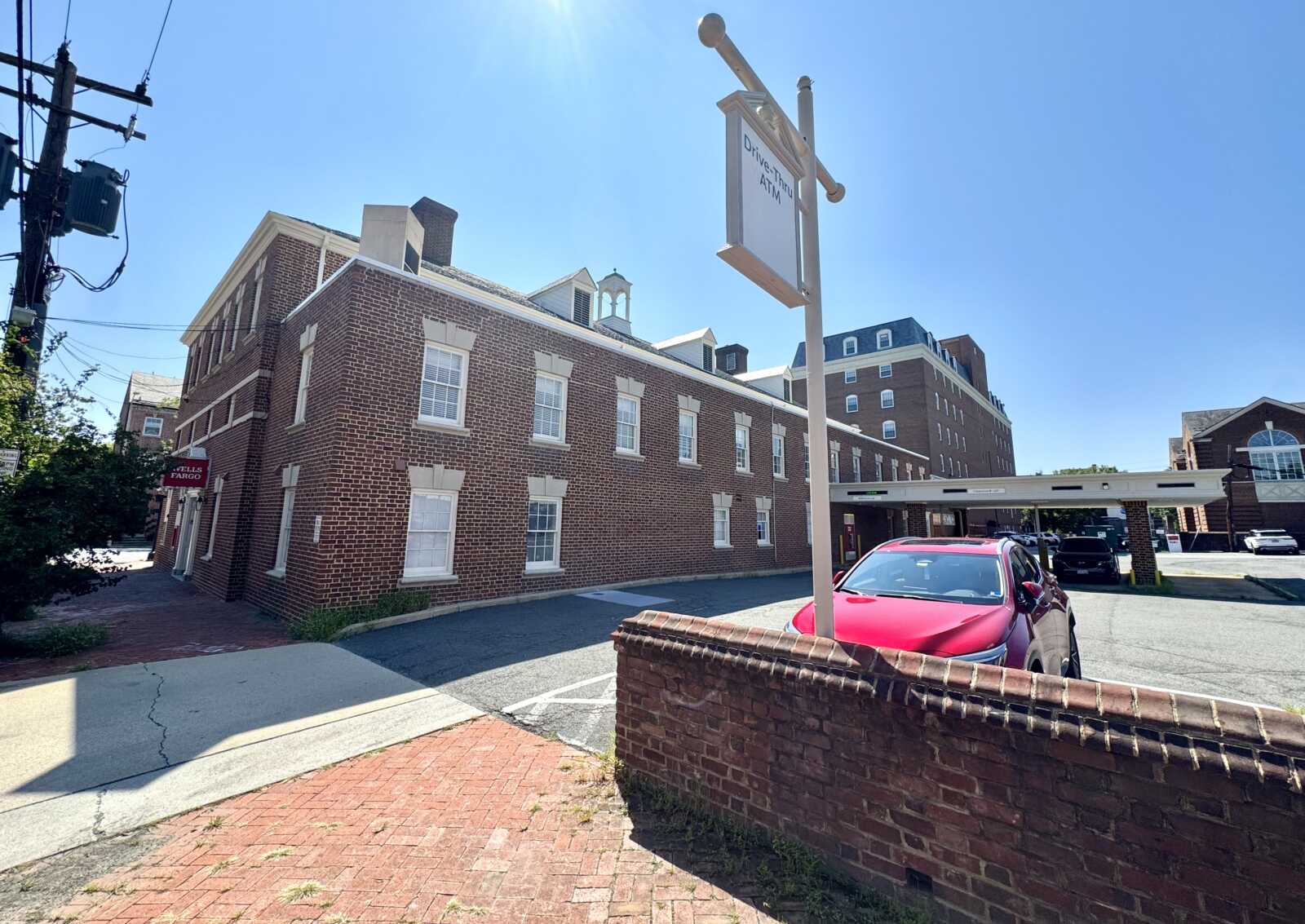 Brick building on a sunny street with a drive-thru ATM sign and a bright pink car in front.