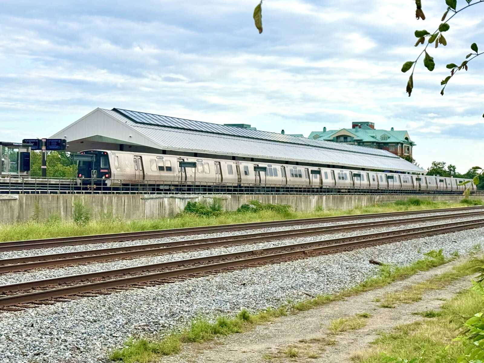 Silver commuter train on an elevated platform with a long white roof, rails and gravel in the foreground, and a cloudy sky above.