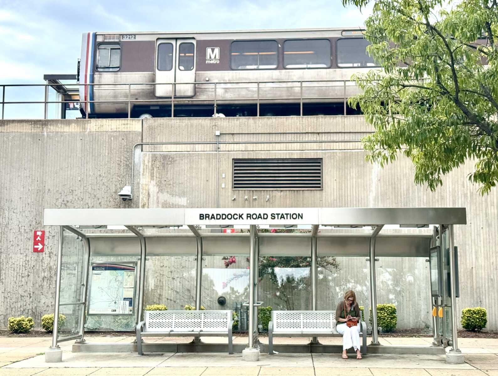 IMG_5204 | ALXnow Braddock Road Station entrance with a modern glass shelter in the foreground and a paused elevated metro car on the track above, a woman sits at the bench using her phone.