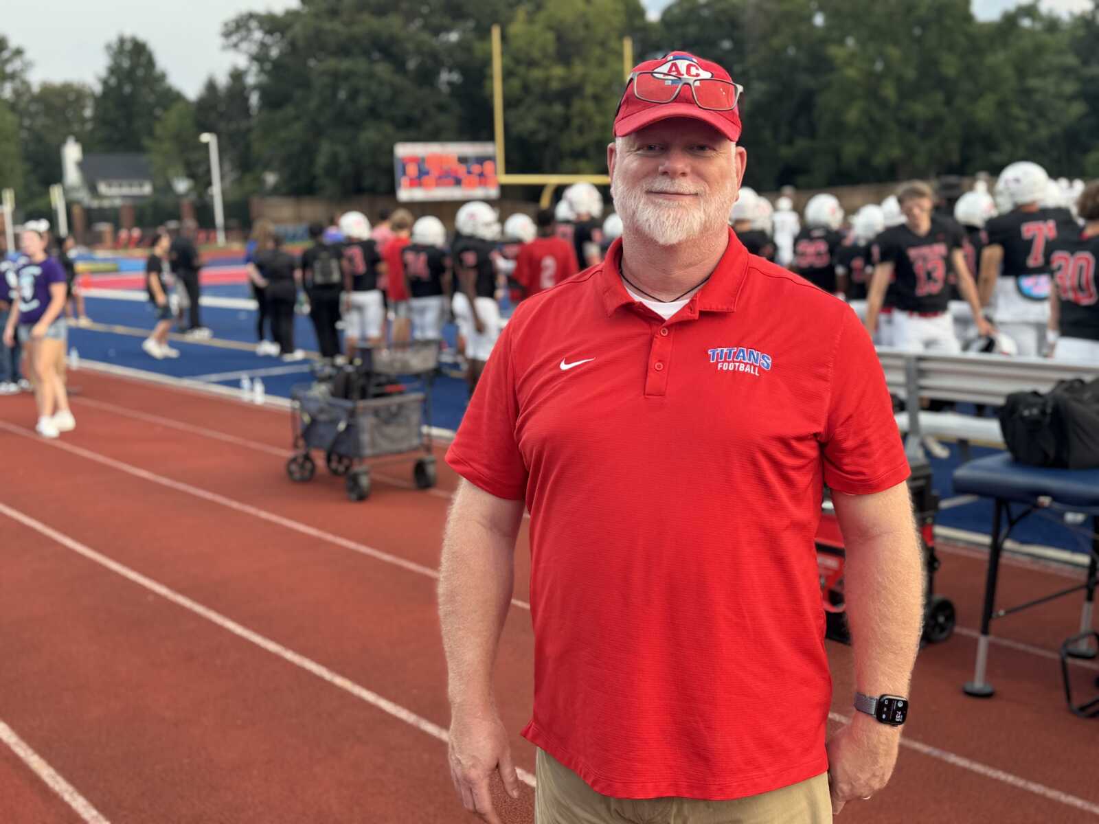 Smiling coach in a red Titans Football polo stands on a track with players on the football field behind him.