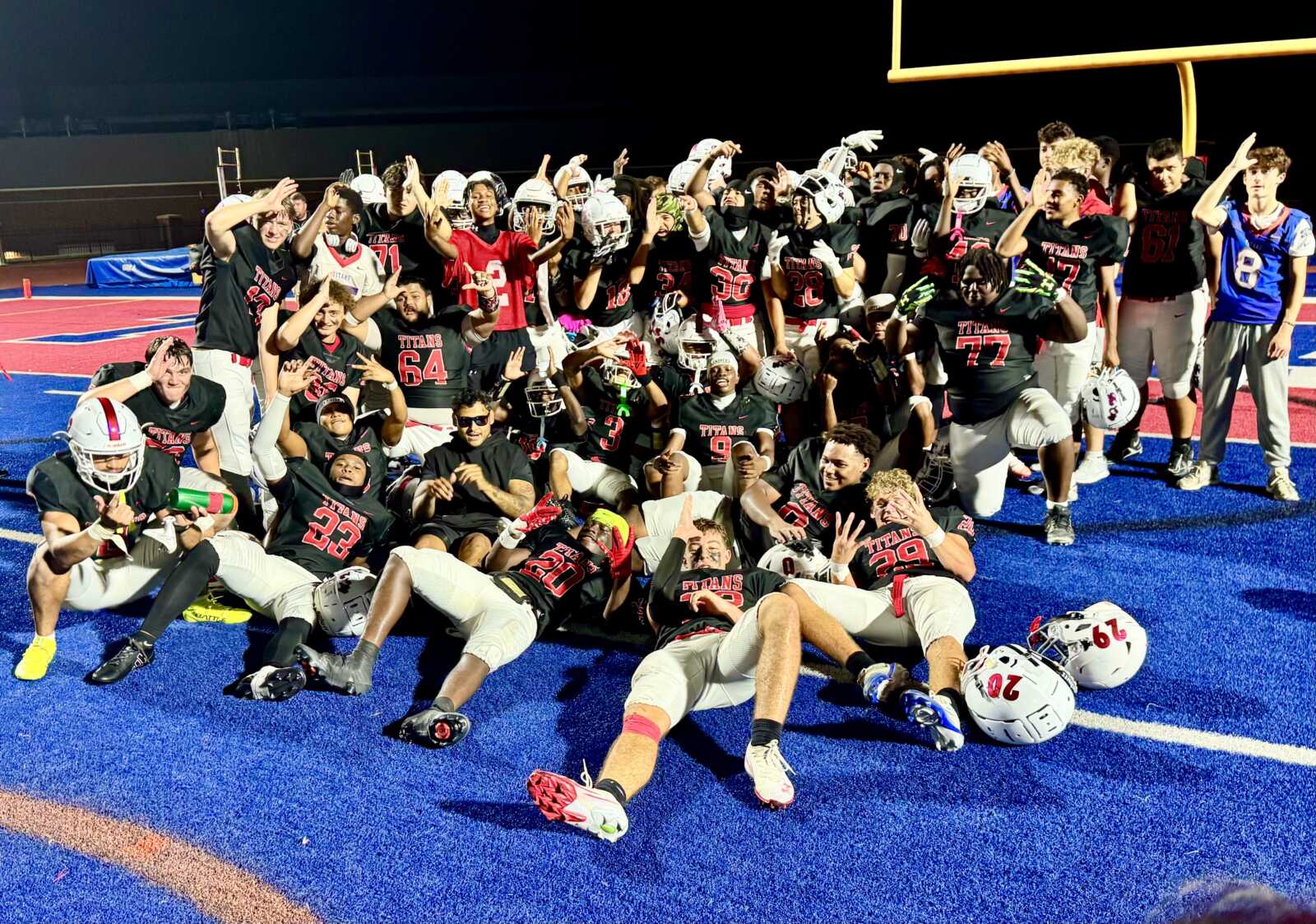 Team of football players and coaches celebrate on a blue turf field, many raising their arms and making victory gestures after a game; helmets on the ground nearby.