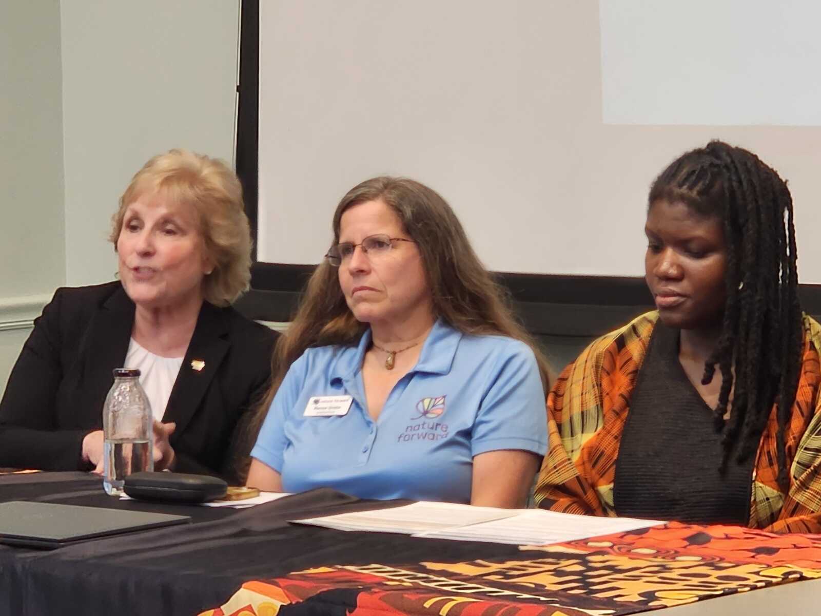 Three women sit at a panel table; left speaks, middle in a blue Nature Forward polo looks on, right woman with braids and patterned wrap listens.