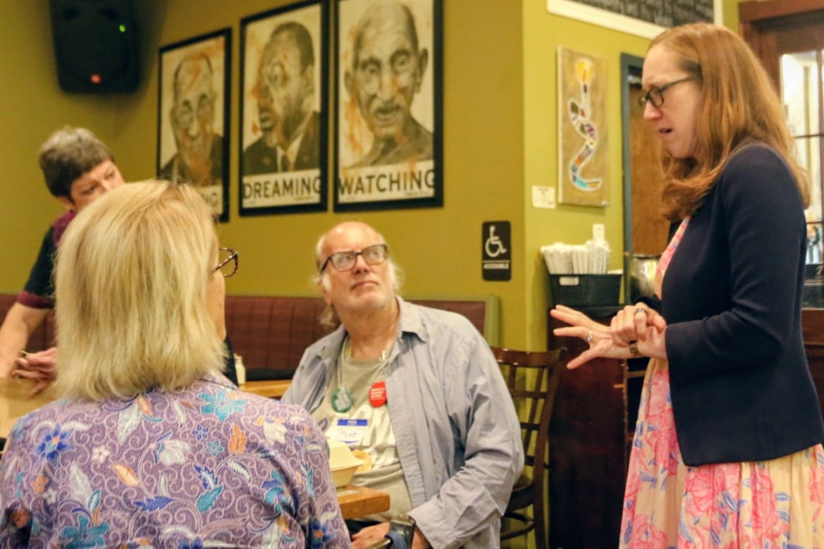 Woman standing and speaking to an older man seated at a table in a cafe; two other people listen in the background with green walls and framed posters behind them.