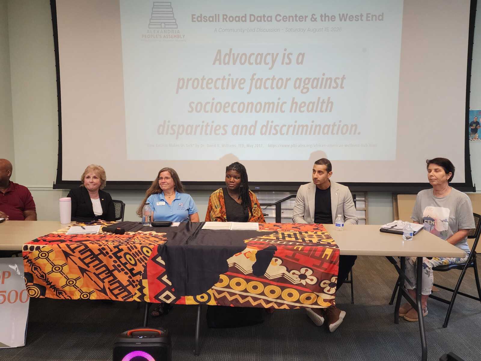 Six panelists seated at a long table in a conference room, with a colorful cloth covering the table and a projection screen behind them.