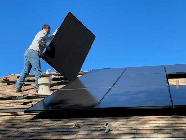 Workman on a pitched roof securing a large black solar panel against a clear blue sky.