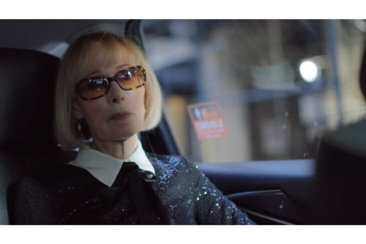 Older woman with blonde bob in sunglasses sits in the back seat of a car, wearing a sequined outfit and white collar.