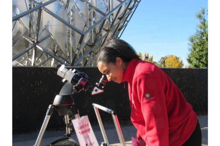 Person in a red jacket leans in to look through a telescope at an outdoor science exhibit, with a metal lattice structure in the background.