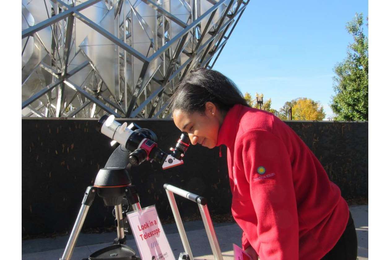 Person in a red jacket leans in to look through a telescope at an outdoor science exhibit, with a metal lattice structure in the background.