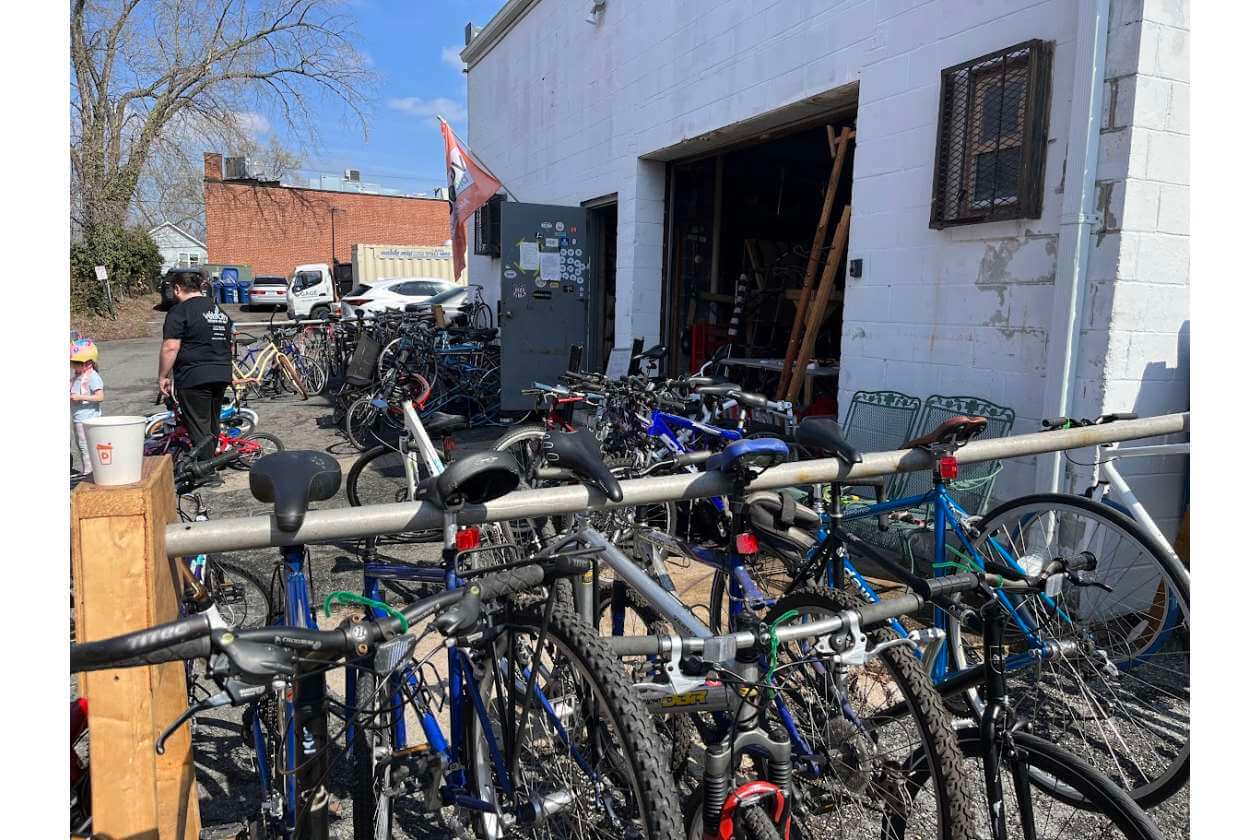 Crowded bike rack in an outdoor lot outside a white building with an open garage, many bicycles chained and parked nearby.