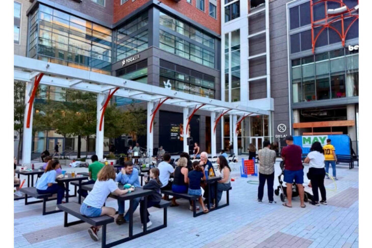 Outdoor city plaza with people sitting at long picnic tables, chatting and playing in front of glass-fronted buildings and a YogaSix storefront in the background.