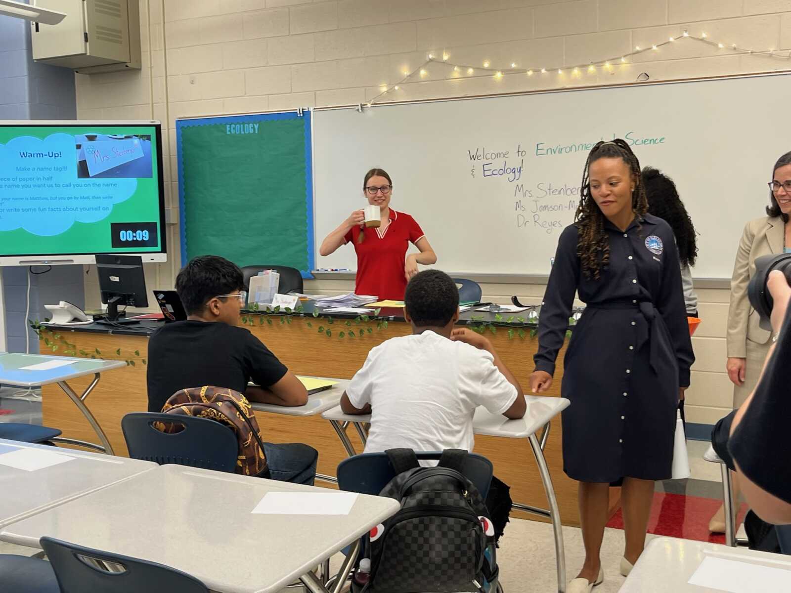 Science classroom scene with a teacher in a red shirt at the front holding a mug, students seated at desks, and a woman in a dark dress standing nearby; whiteboard reads 'Welcome to Environmental Science & Ecology'.