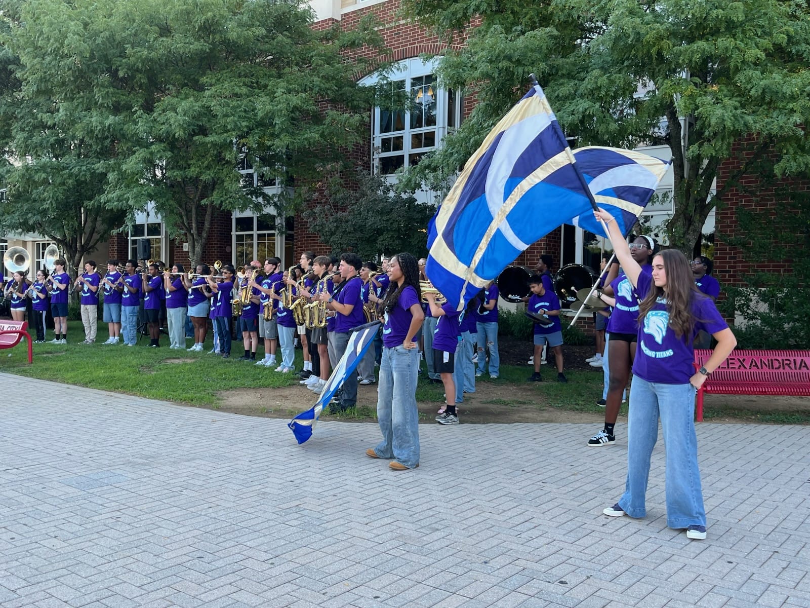 School marching band in purple shirts performs outdoors in front of a brick building, with students playing brass instruments.