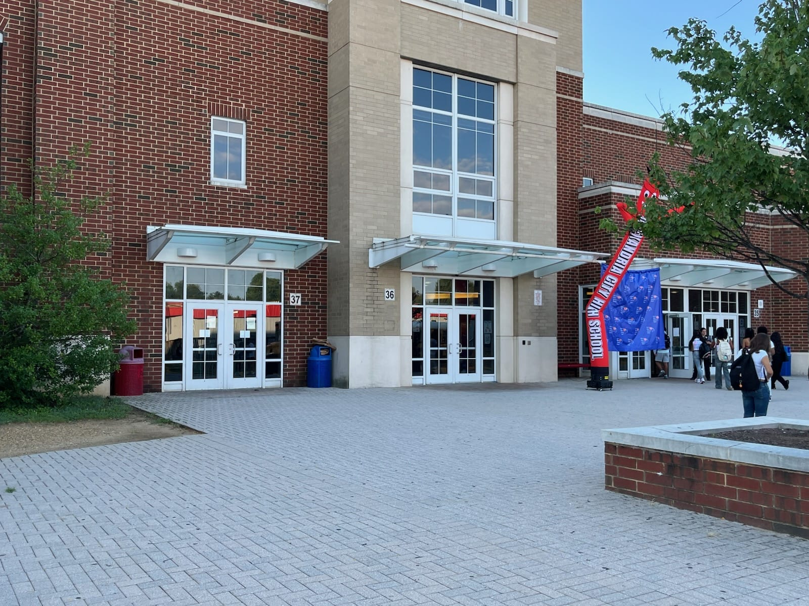 School entrance with glass doors under a metal canopy; brick walls, beige center section, and numbers 36 and 37 above the doors; a blue banner structure and a group of students near the doors on a paved plaza.