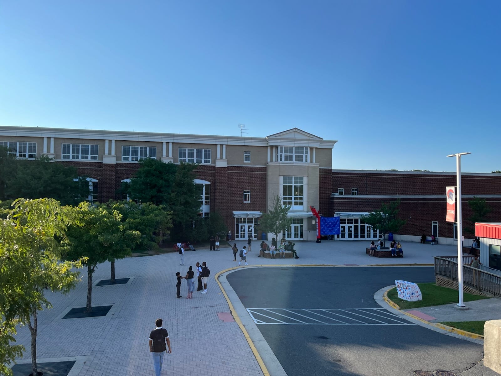 Wide view of a brick school building with a central entrance, surrounding trees, and a paved courtyard where students mingle on a sunny day.