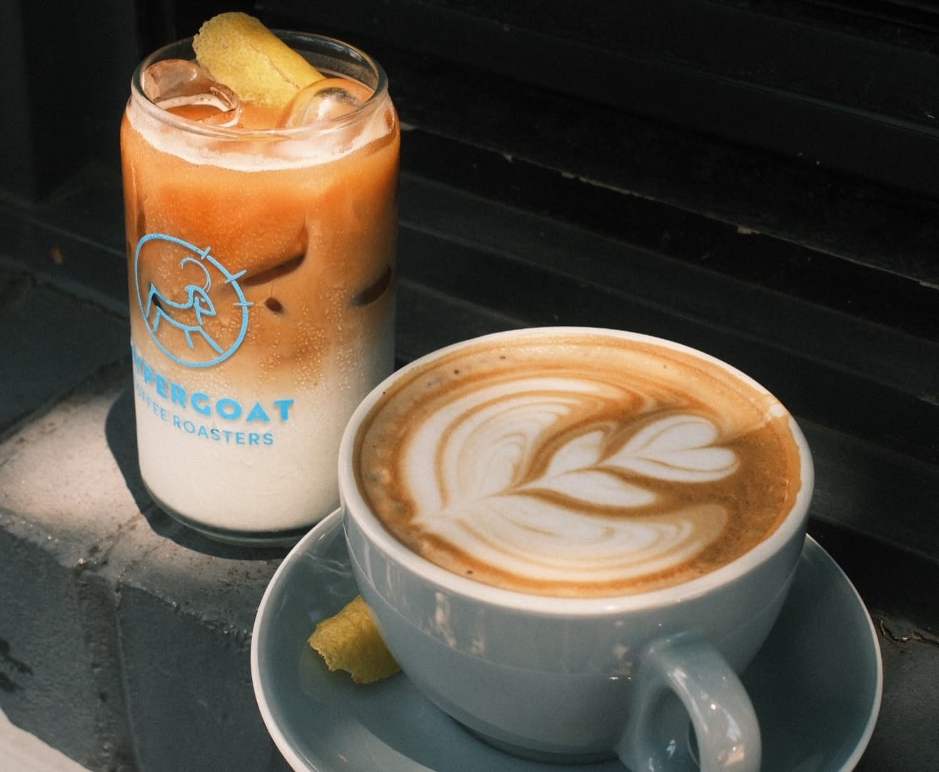 Tall glass of iced coffee with a creamy gradient and a lemon wedge, next to a cup of latte with heart-shaped latte art on a saucer.