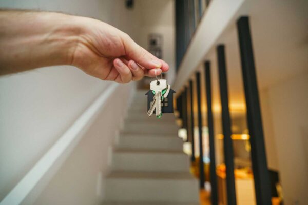 Hand holding a keyring with several house keys and a small house-shaped tag in a residential hallway with a stairwell in the background