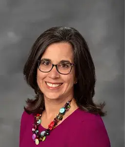 taylor-profile | ALXnow Professional headshot of a smiling woman with dark hair and glasses, wearing a magenta top and colorful beaded necklace.
