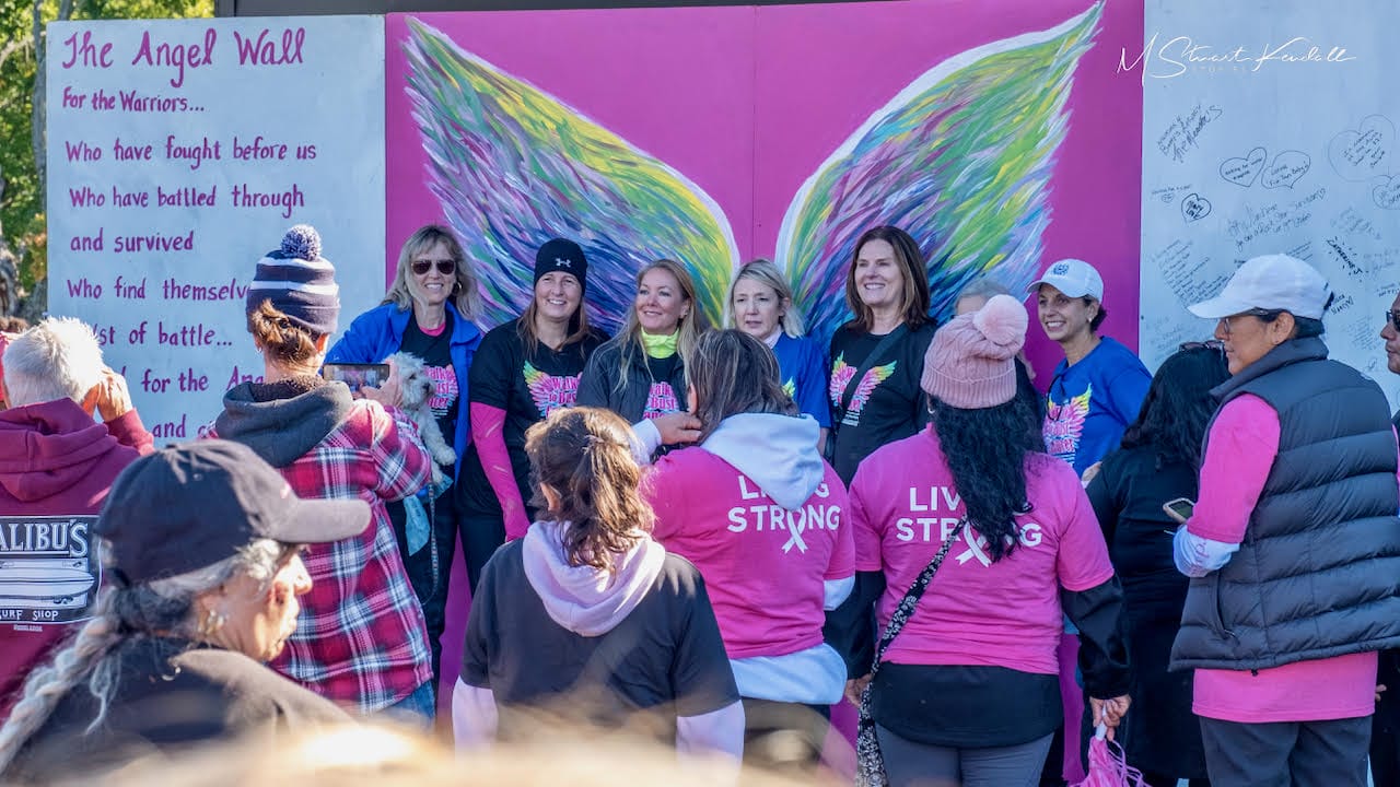 Group of women in pink shirts posing in front of a colorful angel-wing mural and wall text.