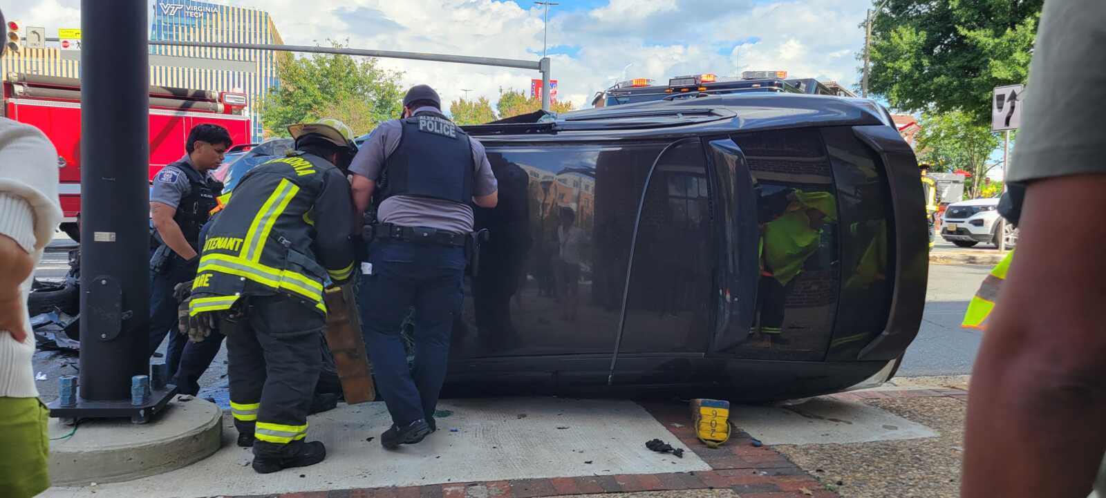 Black SUV on its side at the curb as firefighters and police assist at the crash scene on a sunny urban street, with a fire truck in the background.