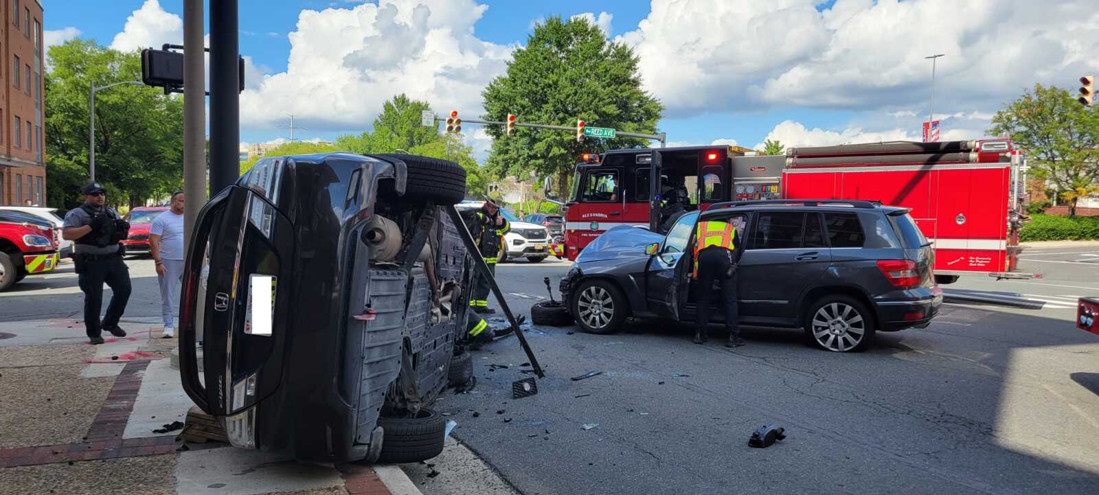 Black SUV lies on its side at an intersection as firefighters and responders work around the scene, with a red fire truck in the background and a gray SUV nearby.