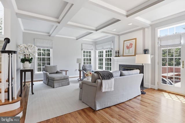 Bright living room with a gray sectional, two armchairs, and a fireplace under a coffered ceiling, filled with natural light from large windows.